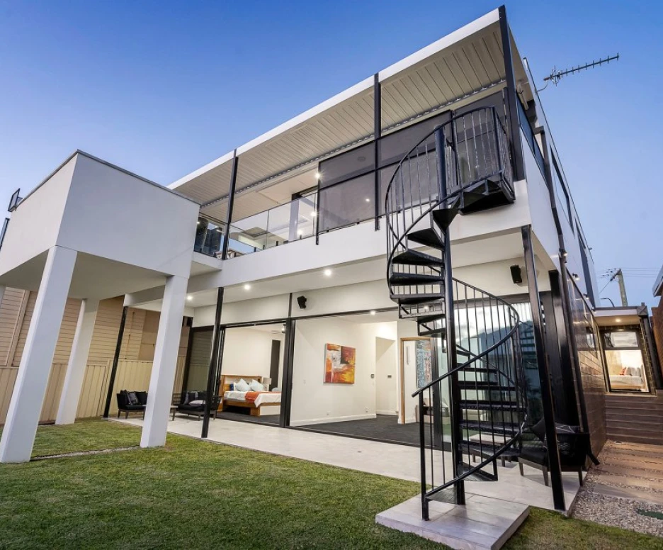 A modern two-story house with a spiral staircase leading to a balcony. The building features large glass windows and a lush green lawn in front. The exterior is a mix of white and beige, with outdoor seating visible.