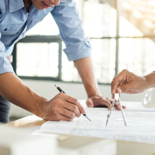 Two people are collaborating on architectural plans, one using a compass and pencil while the other takes notes. A hard hat and construction materials are visible in the background, suggesting a design or construction environment.