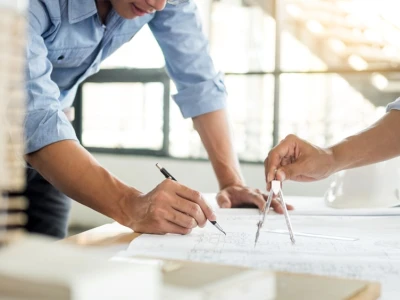 Two people are collaborating on architectural plans at a table, one using a compass while the other sketches with a pencil. A construction helmet and various building materials are visible in the background. Natural light streams in through large windows.
