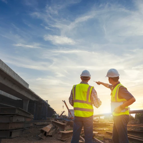 Two construction workers in yellow vests and hard hats stand on a construction site, discussing plans against a backdrop of a vibrant sunset and partly cloudy sky.
