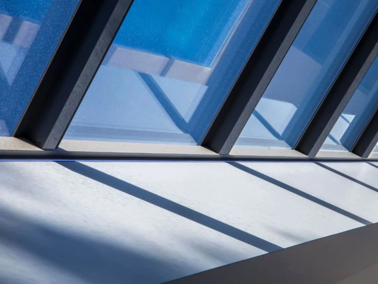 Sunlight filters through a series of angled windows, casting geometric shadows on a white surface below. The blue sky is reflected in the glass, creating a tranquil and modern aesthetic.