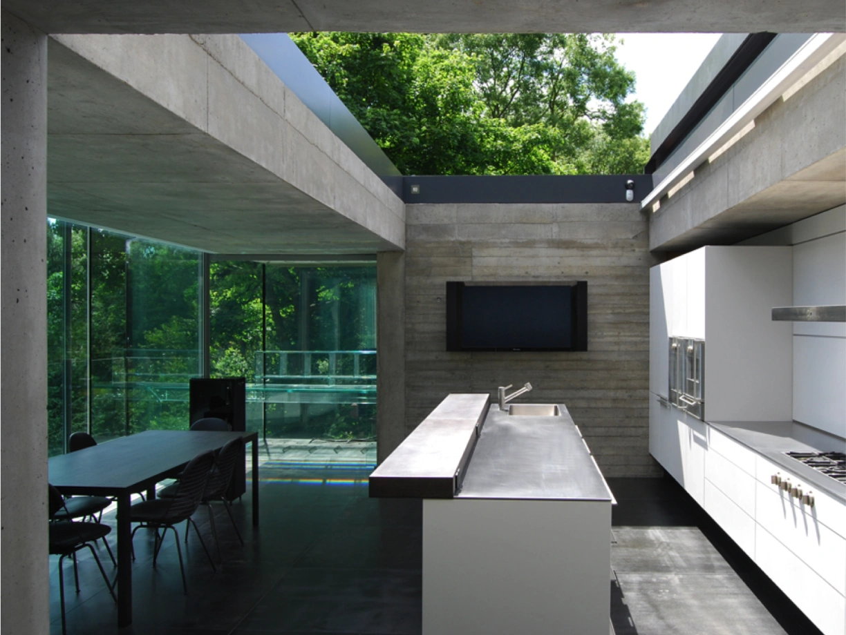 Intérieur d'une cuisine moderne avec des murs en béton et de grandes fenêtres. Un plan de travail en acier inoxydable et une table à manger sont visibles. La lumière naturelle entre par le plafond en verre, entouré d'une verdure luxuriante.