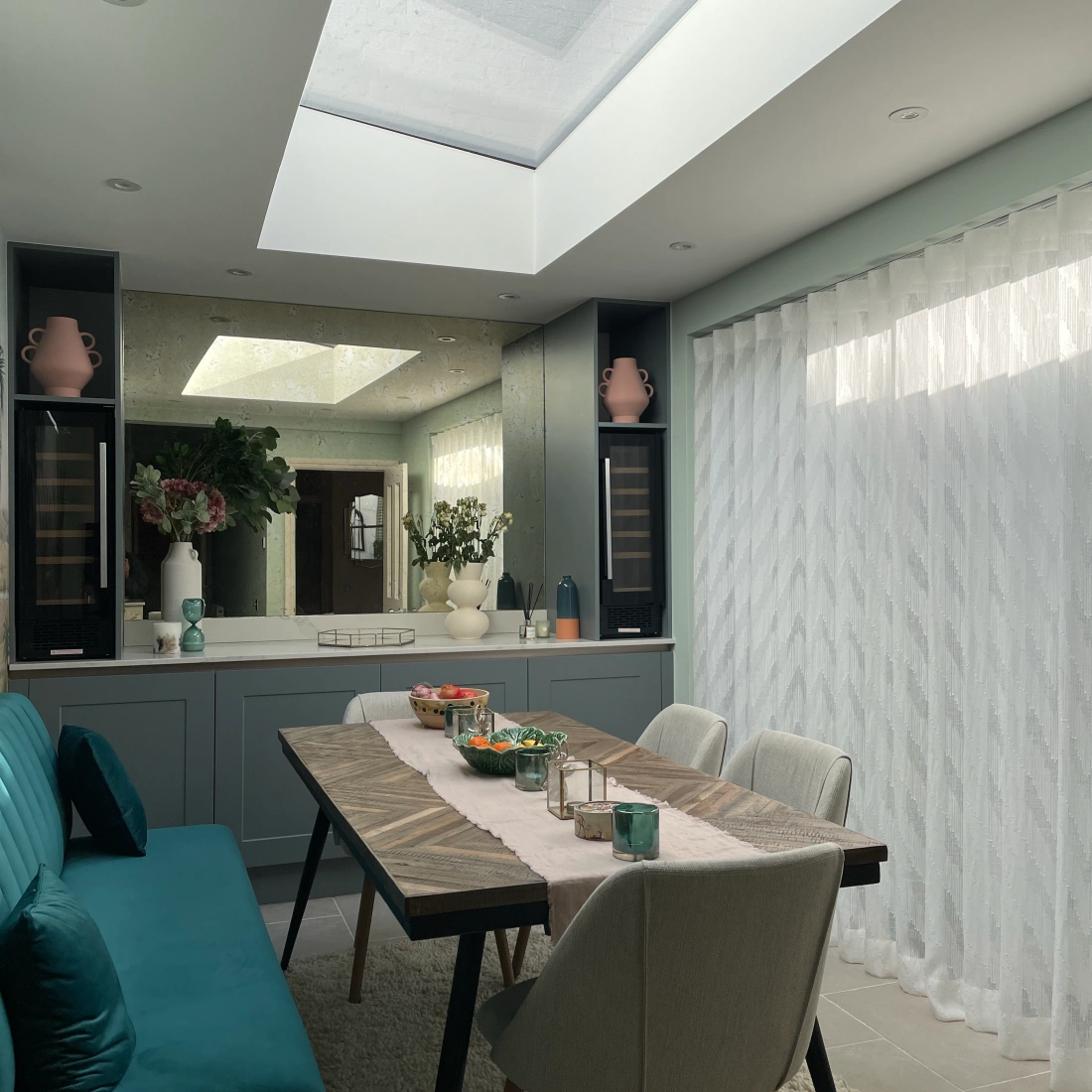 Une salle à manger moderne avec une table au centre, entourée de chaises. Un plafond en verre laisse entrer la lumière, tandis qu'un canapé turquoise est sur le côté. Des vases et des décorations ajoutent une touche élégante.