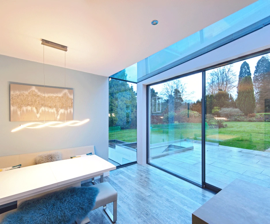 Un intérieur moderne d’une salle à manger avec une grande fenêtre donnant sur un jardin verdoyant. Une table blanche élégante est entourée de sièges confortables. Des lumières suspendues ajoutent une ambiance chaleureuse.