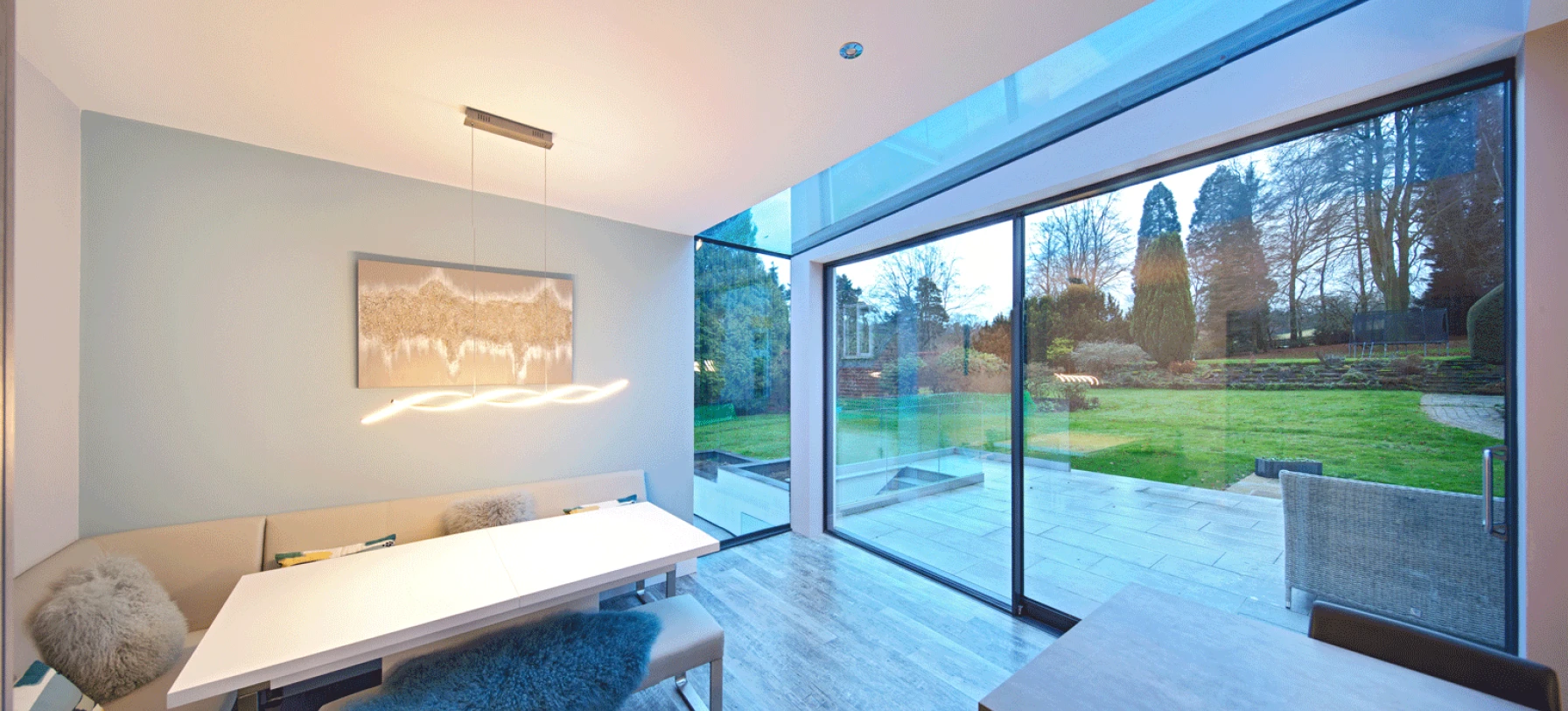Comedor moderno con una mesa blanca y sillas tapizadas. Gran ventana de vidrio que da a un jardín bien cuidado. Iluminación suave y decoraciones minimalistas en tonos neutros.