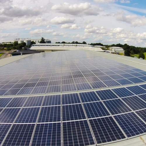 A wide view of a rooftop covered in solar panels, reflecting sunlight under a partly cloudy sky. In the background, residential buildings and green fields are visible.