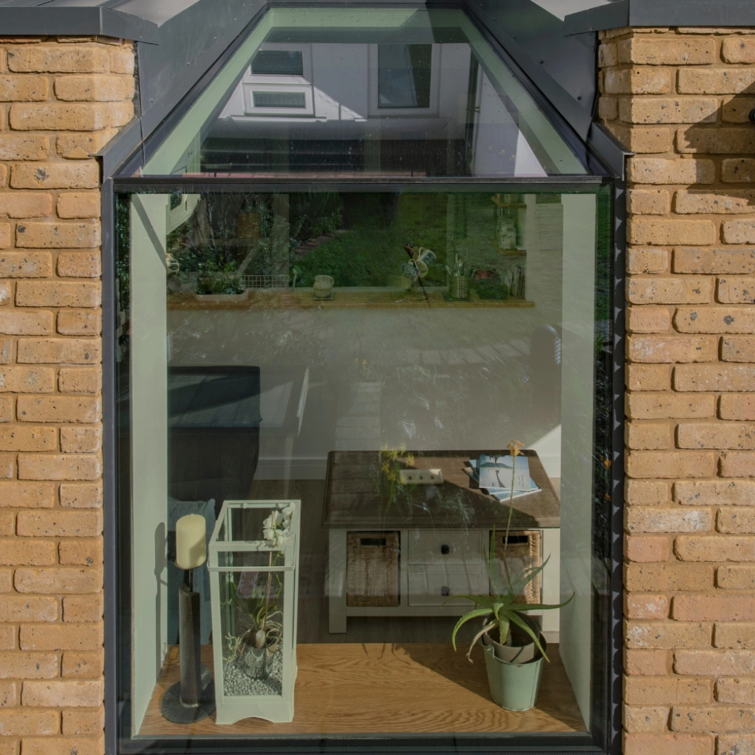 Ventana de una habitación moderna con una mesa de madera, una planta en maceta y un acuario. Se pueden ver detalles del jardín exterior reflejados en el cristal.