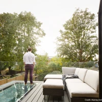 A man stands on a balcony, looking out at trees and a cloudy sky. The balcony features a modern outdoor sofa and a cozy throw blanket.