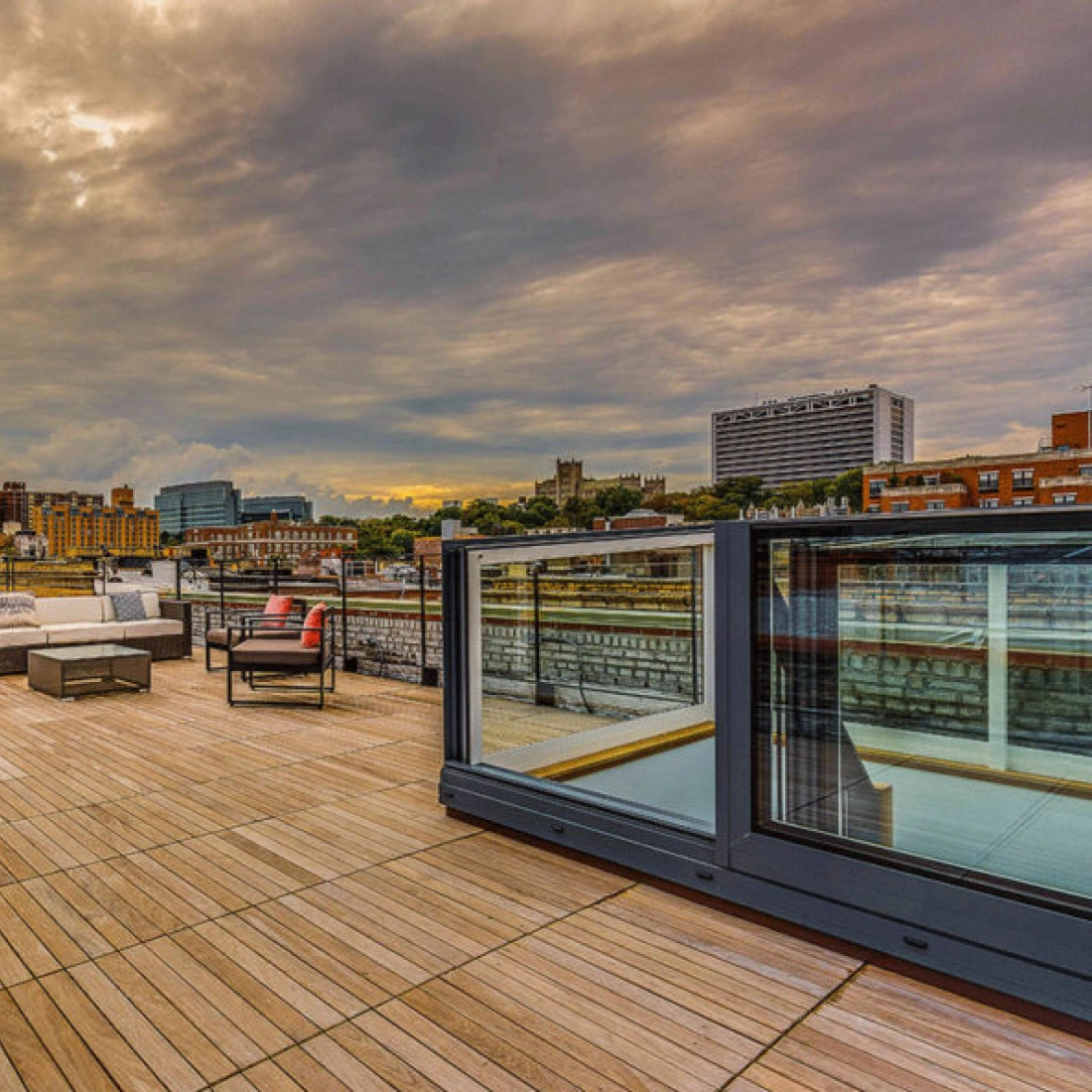 A spacious rooftop terrace featuring wooden flooring, outdoor seating arrangements, and glass railings. The skyline includes buildings and a cloudy sky in the background.