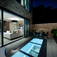 A modern patio area at night features a glass panel floor revealing blue lighting underneath. It opens up to a cozy living room with a light sofa and contemporary decor. Potted plants and a dining table complete the elegant outdoor space.