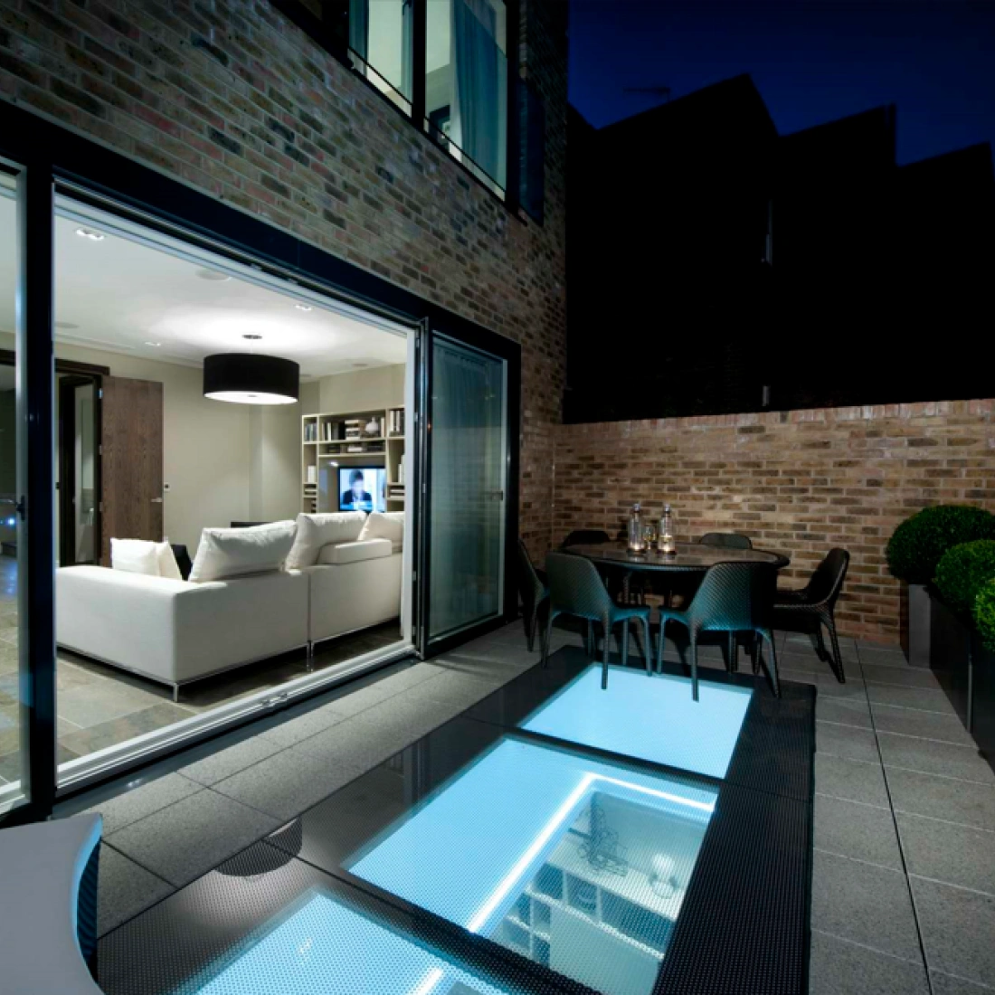 Terraza moderna con piso de vidrio iluminado, mesa y sillas, y una sala de estar visible a través de grandes ventanales. El ambiente es nocturno, con una iluminación suave y un diseño elegante.