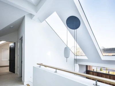 A modern, well-lit hallway with a sloped ceiling and large skylights. A sleek, minimalistic hanging light fixture is suspended from the ceiling. The walls are painted white, and the railing is made of wood.