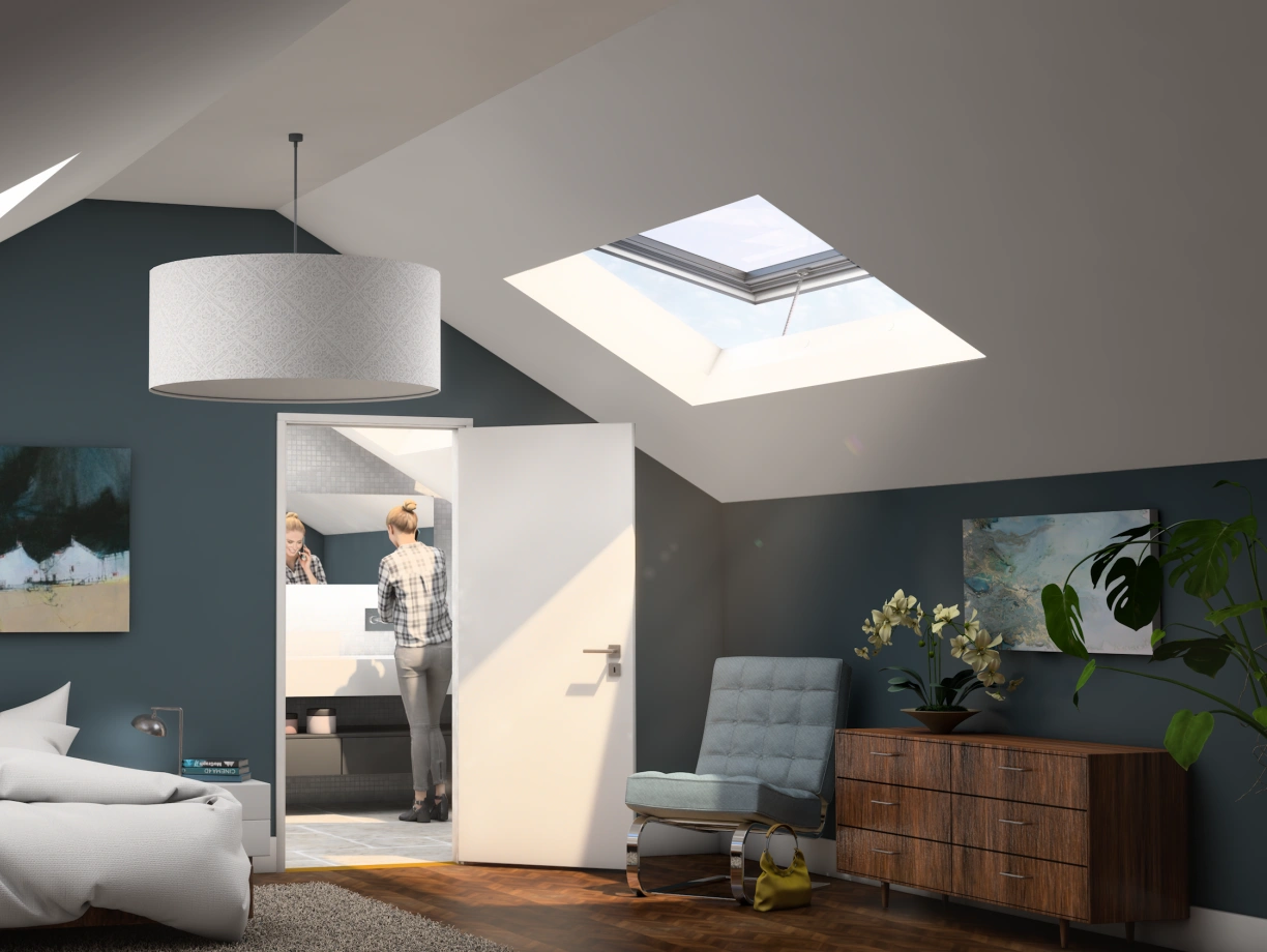 A cozy attic bedroom with a white bed, a hanging light fixture, and dark blue walls. Two paintings are displayed on the walls, and a person is seen in the bathroom beyond the open door. Natural light enters through skylights, illuminating the space. A modern chair and a wooden dresser complete the decor.