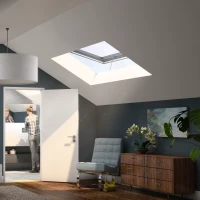 Chambre sous les combles avec un lit en désordre, un plafond en pente, des lucarnes laissant entrer la lumière, une peinture accrochée au mur et une porte menant à une salle de bain où se trouve une personne. Un fauteuil et une commode complètent l'espace.