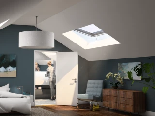 Habitación con un techo inclinado, paredes de color azul oscuro y grandes ventanales en el techo. Hay una cama deshecha, una silla moderna gris y un mueble de madera. Se ve a una persona en un baño al fondo. Decoración artística en las paredes.