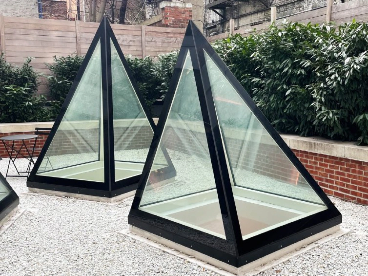 Two modern glass pyramids with black frames sit in a gravel garden area, surrounded by greenery and brick walls. In the background, brick buildings with fire escapes are visible.