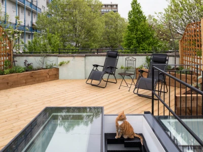 A rooftop garden featuring wooden decking, two black lounge chairs, a small table, and a cat sitting near a set of stairs that lead to a lower level. Lush greenery surrounds the area with buildings in the background.