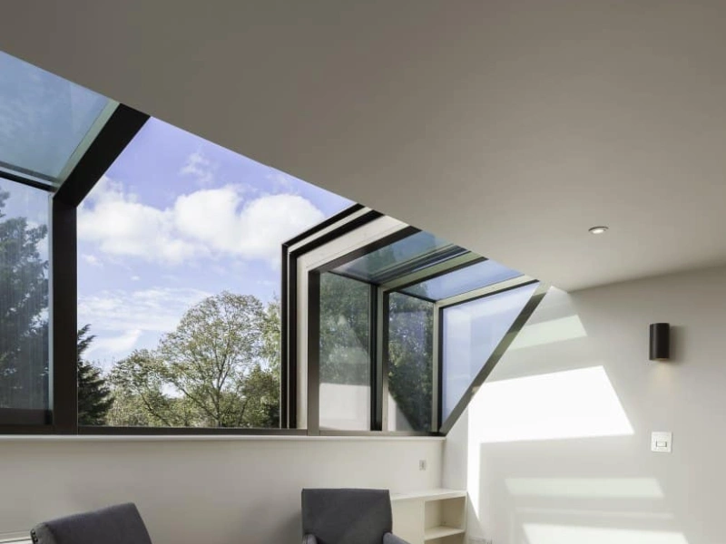 Two blue armchairs with wooden legs sit on a light wooden floor in a bright room. Large windows with a modern design allow natural light to fill the space, showcasing a view of trees and a partly cloudy sky. A built-in shelf is positioned against the wall.