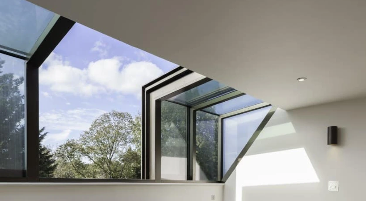 Dos sillones azules en un interior moderno con suelo de madera. Ventanas grandes permiten la entrada de luz natural y ofrecen vistas de árboles exteriores. Las paredes son de un color claro y hay una estantería simple.
