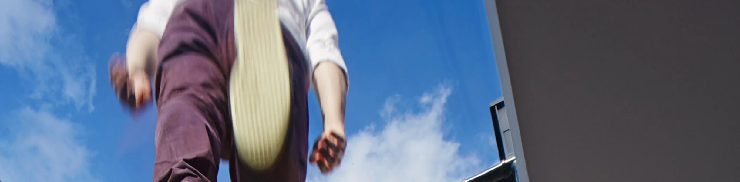Una persona con pantalones oscuros y zapatos claros parece estar caminando sobre un tragaluz, con una vista del cielo azul y nubes. La pared de ladrillo expuesta de una casa se ve detrás.