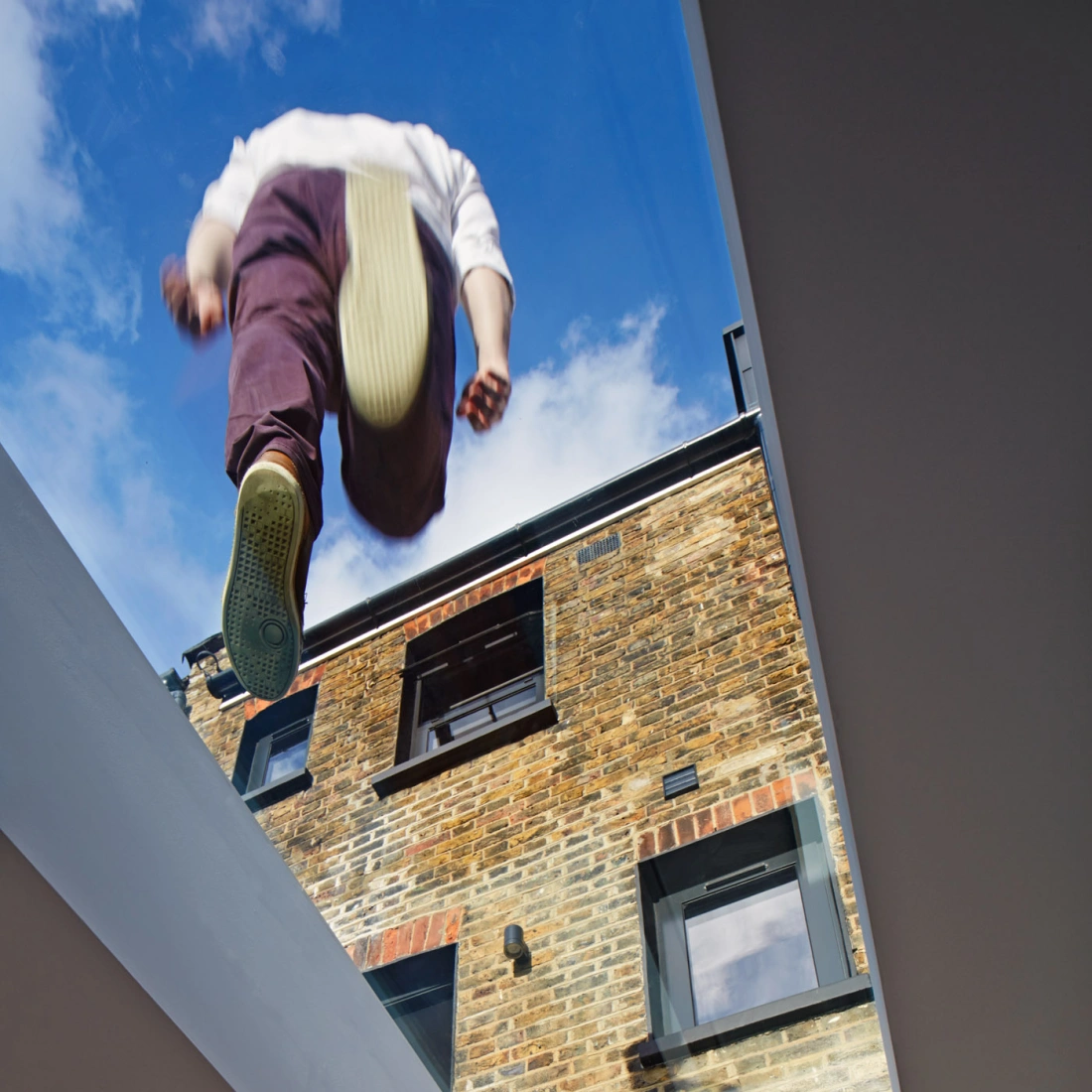Eine Person springt von einer erhöhten Position auf ein Gebäude mit Backsteinfassade. Der Blick ist von unten durch ein Glasdach gerichtet, das den Himmel sichtbar macht.