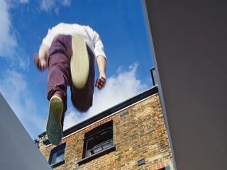 A person is seen walking on a glass skylight, with their feet visible above a brick building. The blue sky peeks through the skylight.