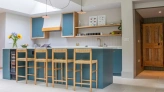 A modern kitchen featuring a large island with four wooden bar stools. The island has a sink and is surrounded by light blue cabinets. Sunflowers are displayed in a vase on the countertop, with a wooden door visible in the background. Natural light floods the space through a skylight.