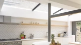 A modern kitchen featuring a gray range cooker, herringbone tiled walls, and a light-filled space with open shelving. A countertop displays a wooden tray with fruits, and large windows provide ample natural light.